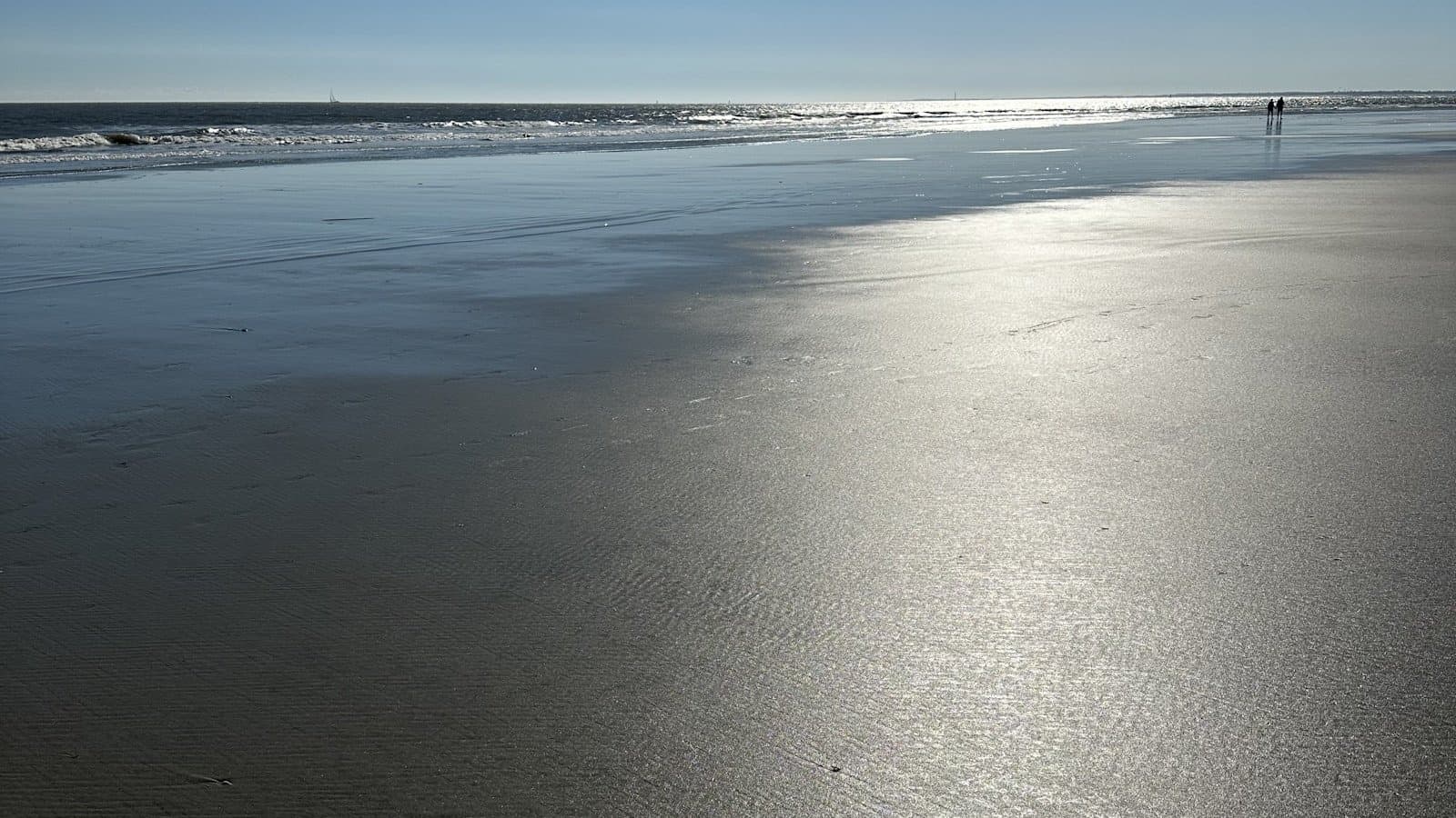 A wide, glassy low-tide beach at Sullivan's Island with two distant figures under a clear sky.
