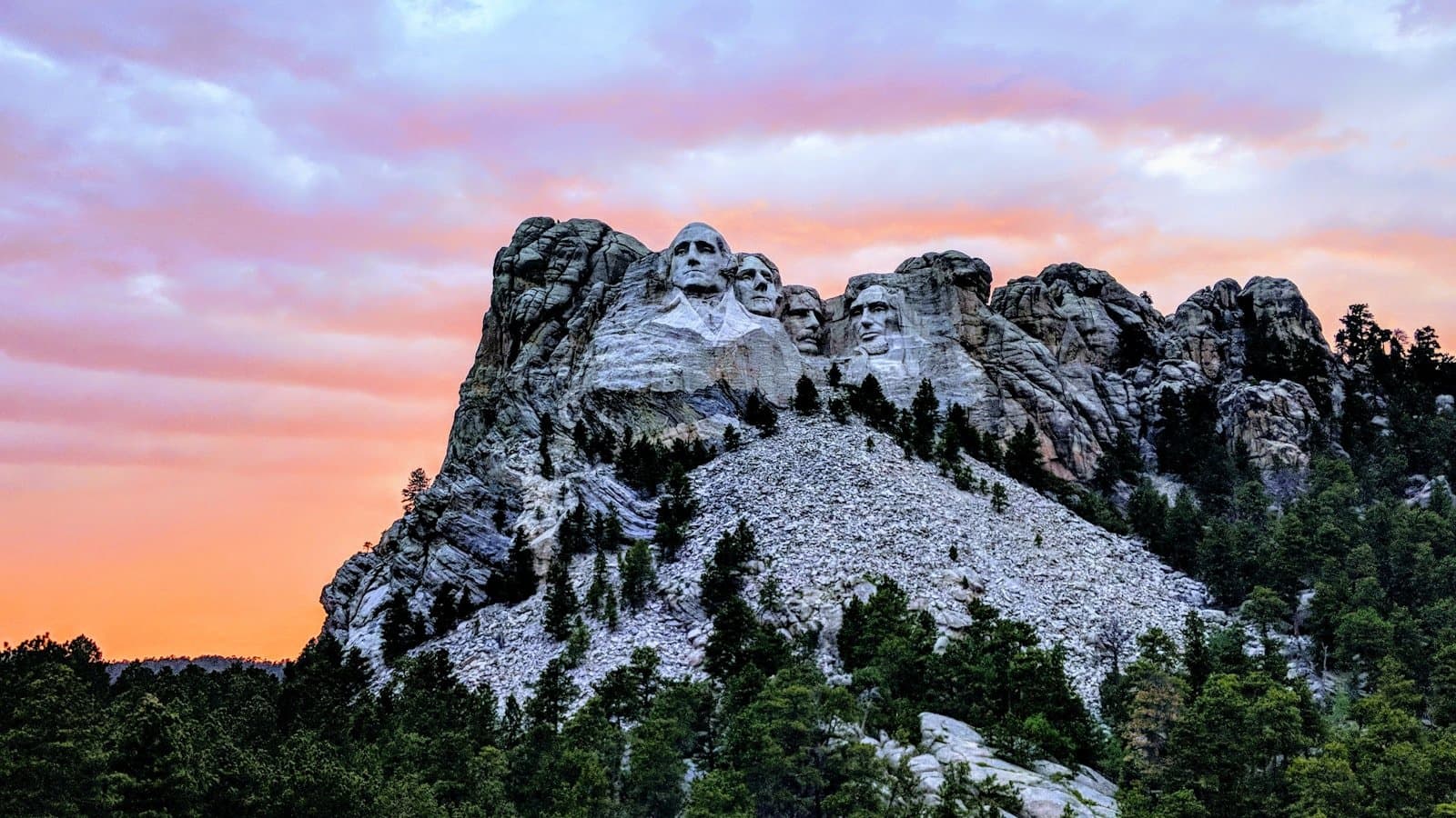 Mount Rushmore National Memorial at sunset, the four carved presidential faces lit against a pink-and-orange sky.