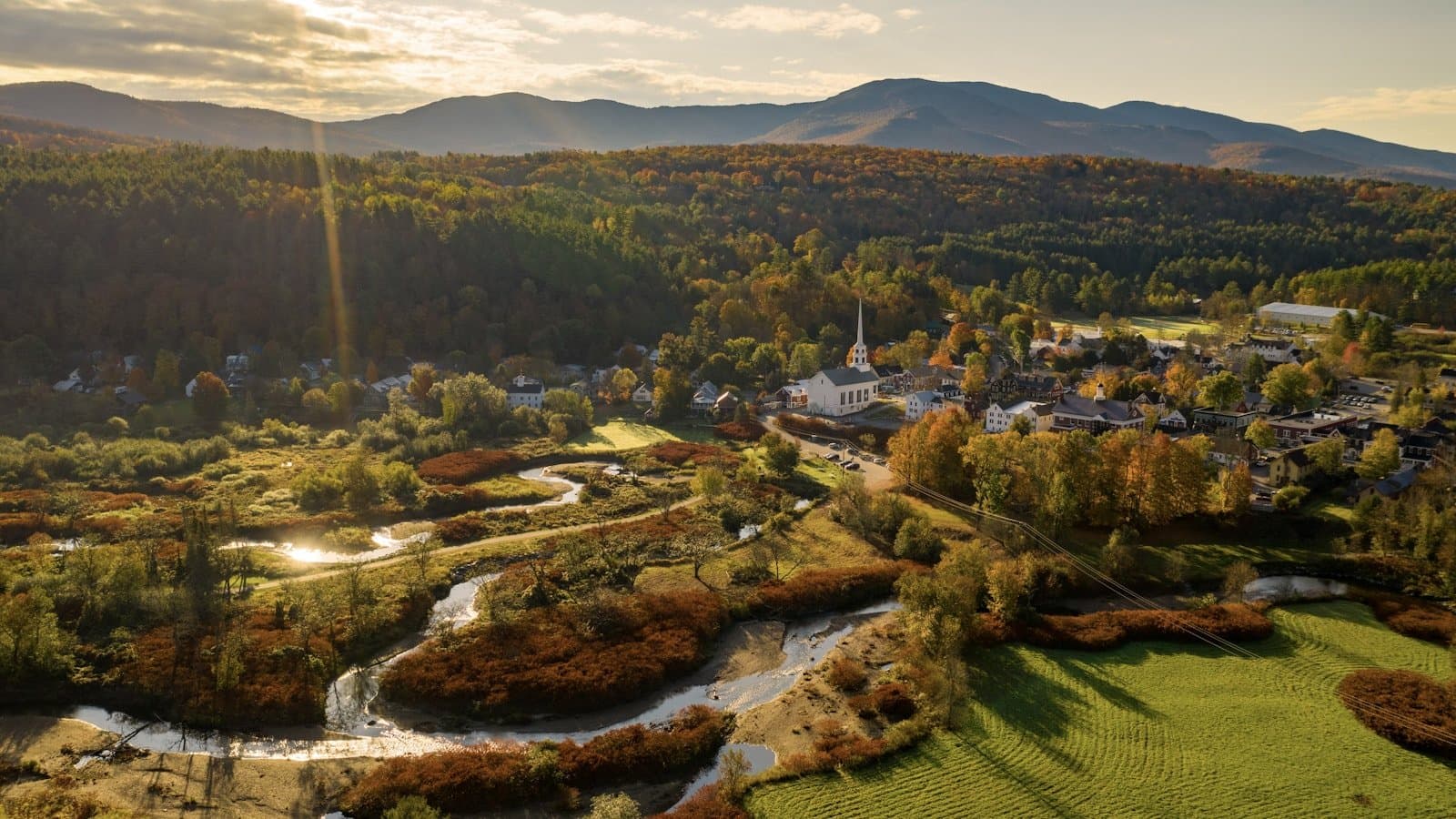 A golden-hour aerial view of the town of Stowe, Vermont, showing its white church steeple, a winding river, and forested hills in fall foliage.