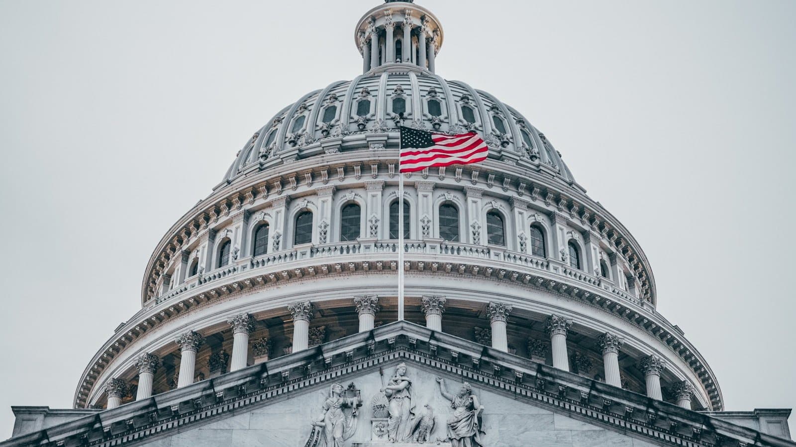 A low-angle view of the U.S. Capitol dome with an American flag flying from its base, columns and pediment sculpture visible below.
