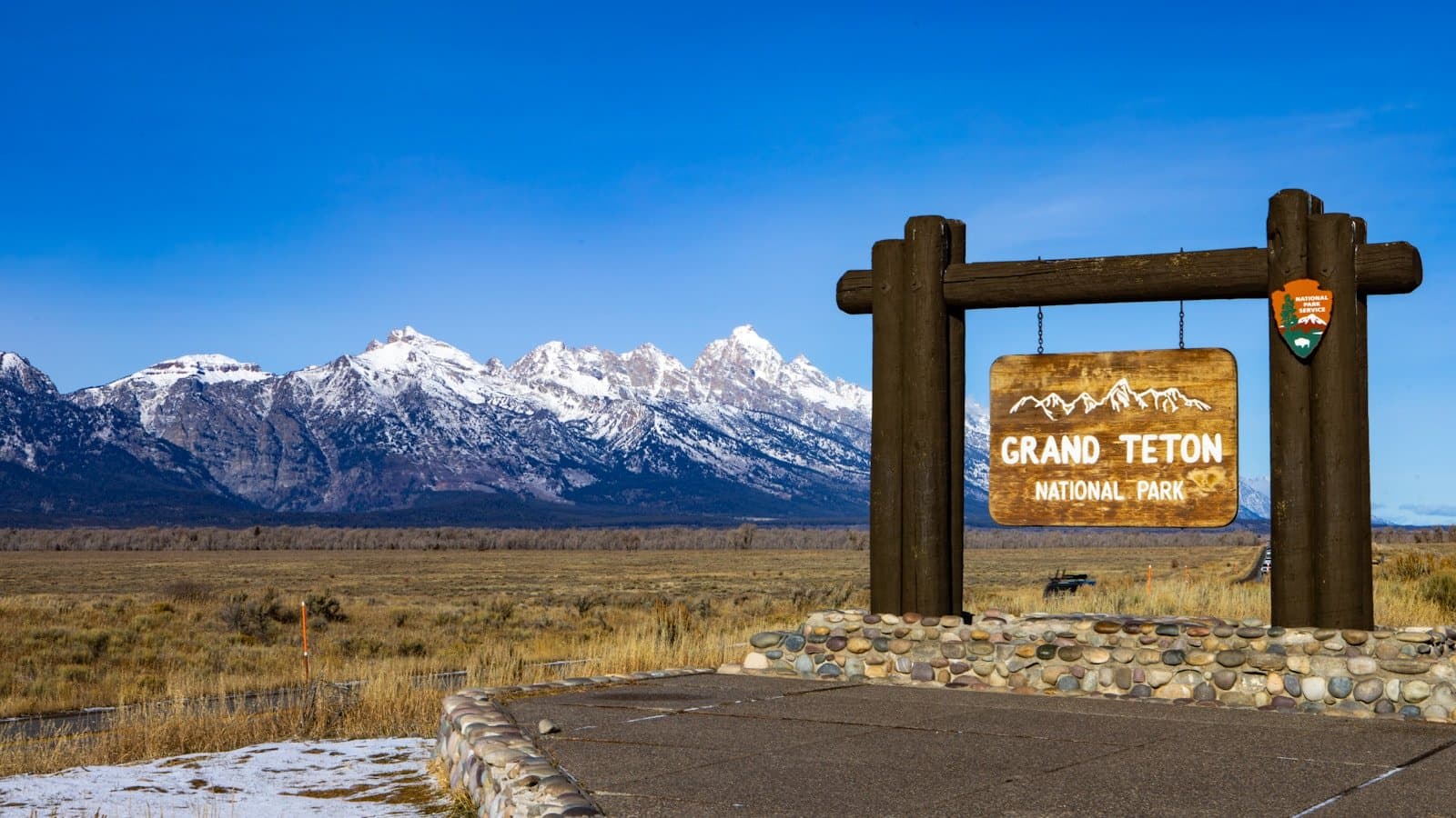 A wooden "Grand Teton National Park" entrance sign in front of the snow-capped Teton mountain range.