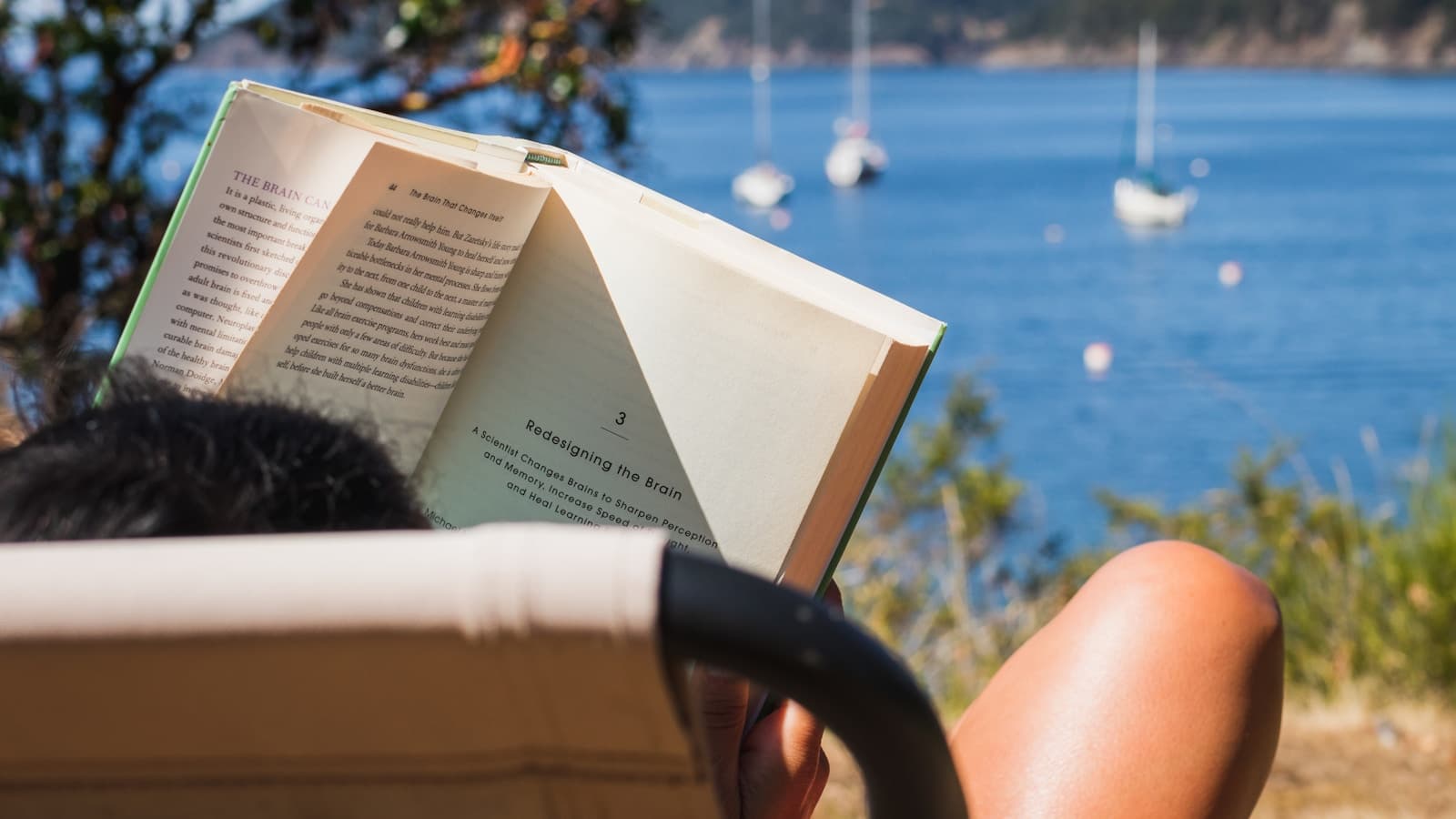 A person reading a book in a beach chair with sailboats in the distance.