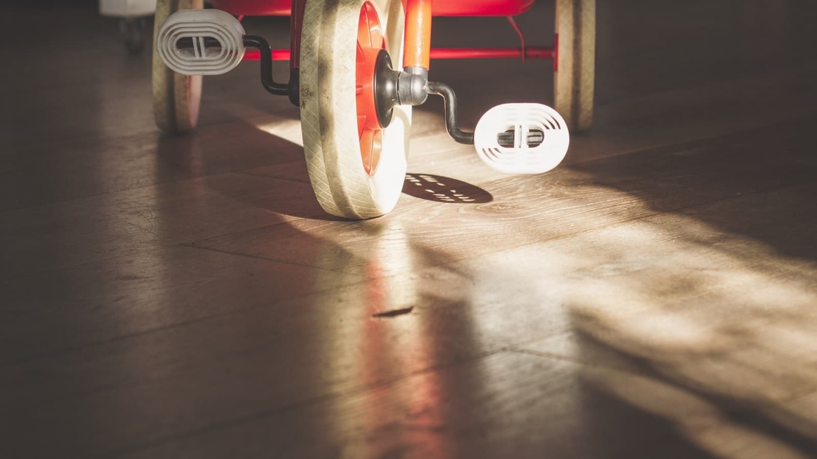 A close-up of a red toddler tricycle's wheels and training wheels on a sunlit hardwood floor, no people, text, or logos.