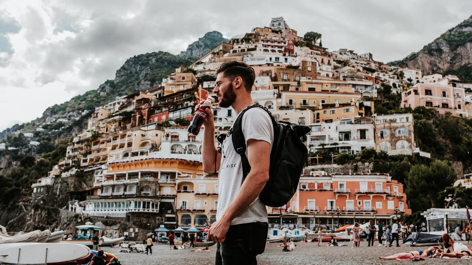 A young man drinking coffee on a colorful hillside coastal town street, backpack on.