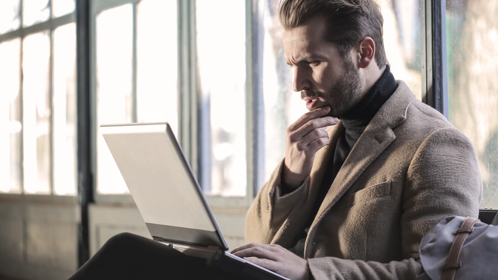 A man in a coat thoughtfully looking at his laptop, hand on chin.