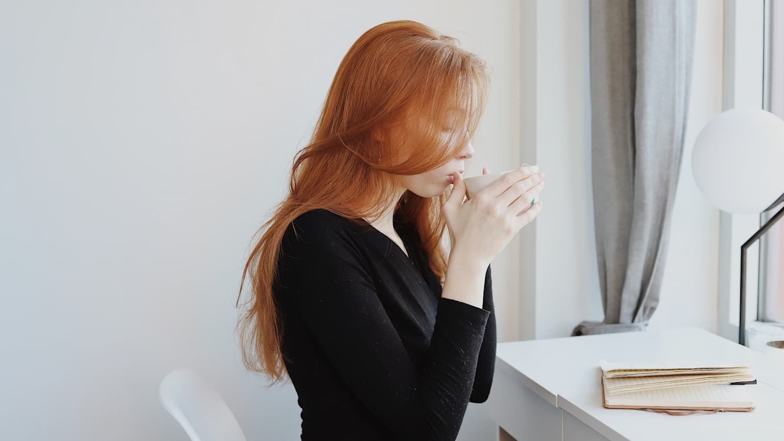 A woman sitting quietly at a desk by a window, pensive expression.
