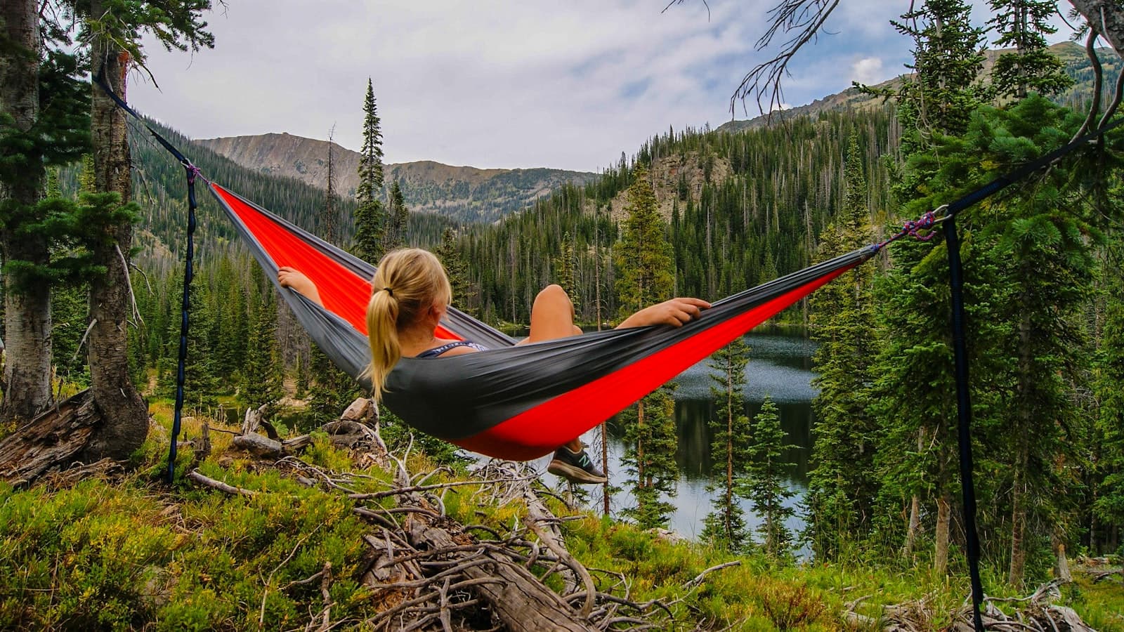 A person relaxing in a red hammock overlooking a mountain lake, seen only from the back of the head; no face or logos.