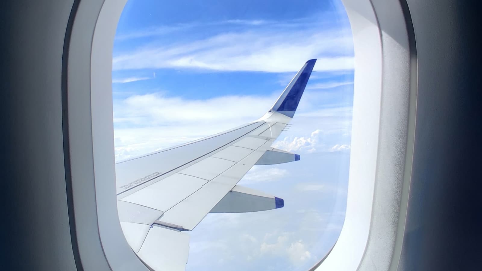 View of an airplane wing against blue sky and clouds through an oval window, with no visible airline branding, text, or people.
