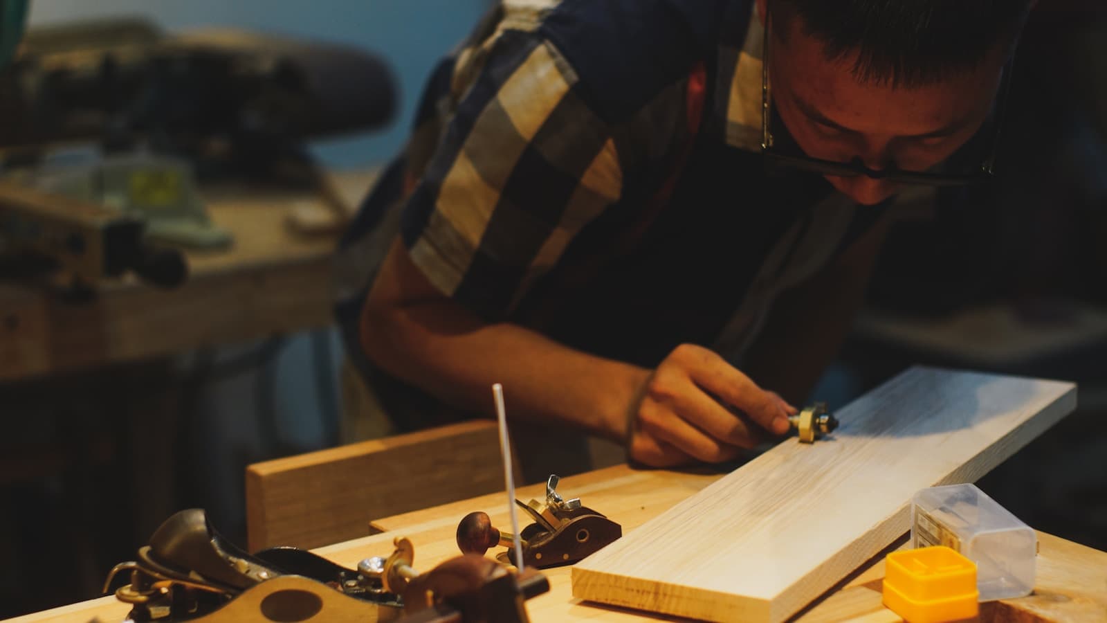 A woodworker bent over a workbench shaping a wood plank with hand tools.