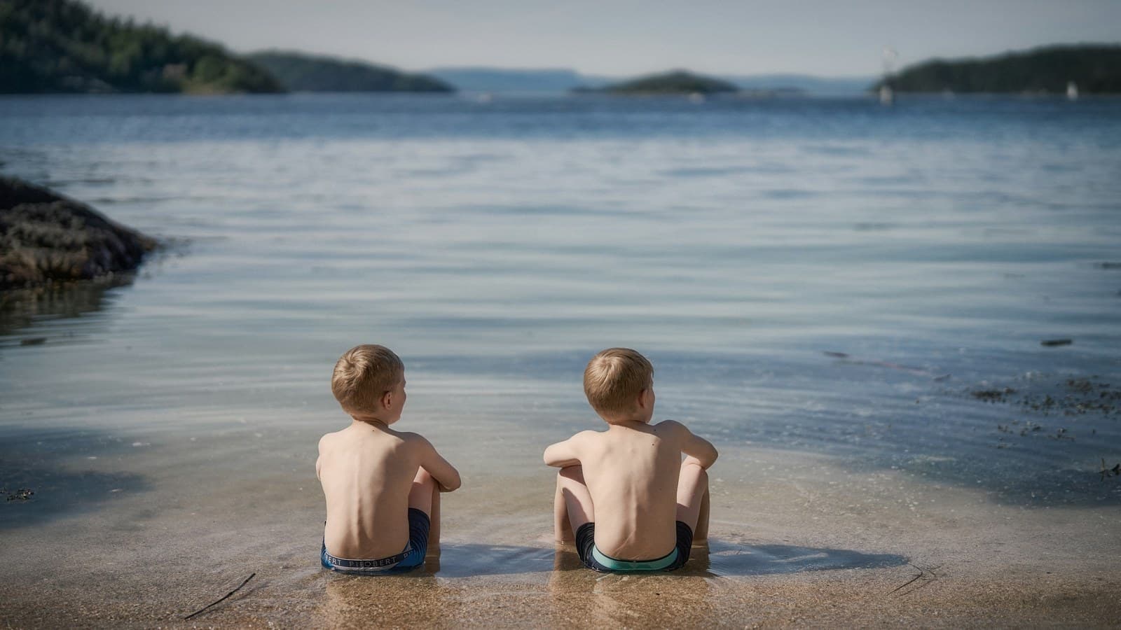 Two young boys sitting together in shallow water at the beach.