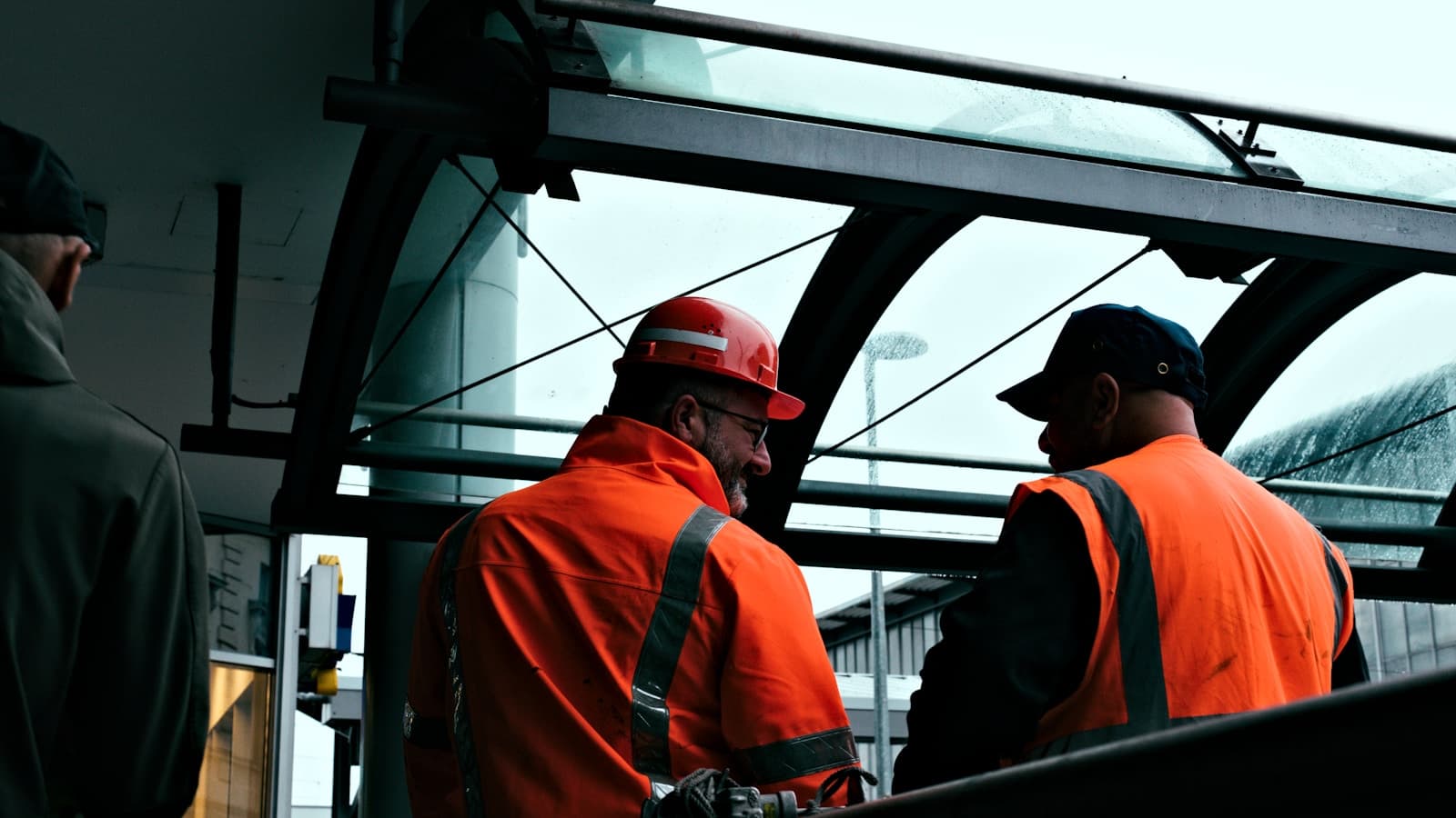Two workers in orange hi-vis vests and hardhats talking under an industrial canopy.