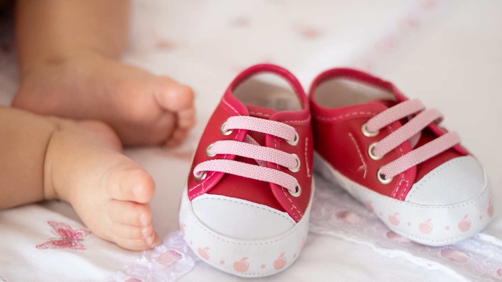 A baby's bare feet next to a pair of small pink sneakers on a bed, no face visible